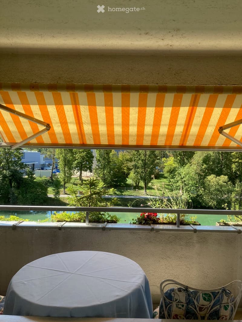 Balcony with a table and chair, covered by an orange and white striped awning, overlooking a green park area.