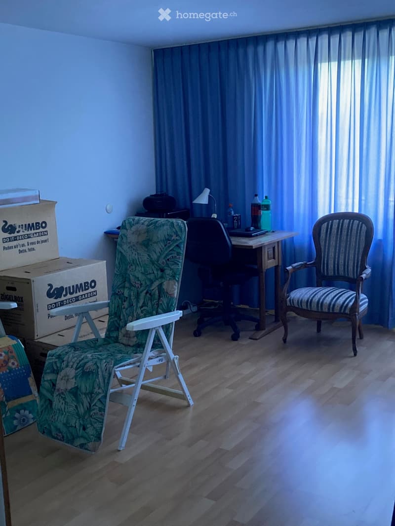 A furnished room with wooden flooring, a large window with blue curtains, a wooden desk with a laptop, a green-covered folding chair, a striped armchair, and several cardboard boxes stacked against the wall.