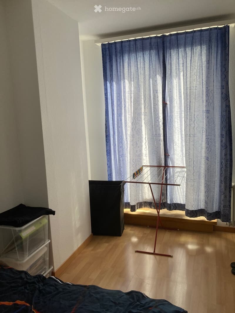 Empty room with wooden flooring, light-colored walls, and a window with blue patterned curtains. A clothes drying rack is positioned near the window, and a small black storage unit is visible on the left side.
