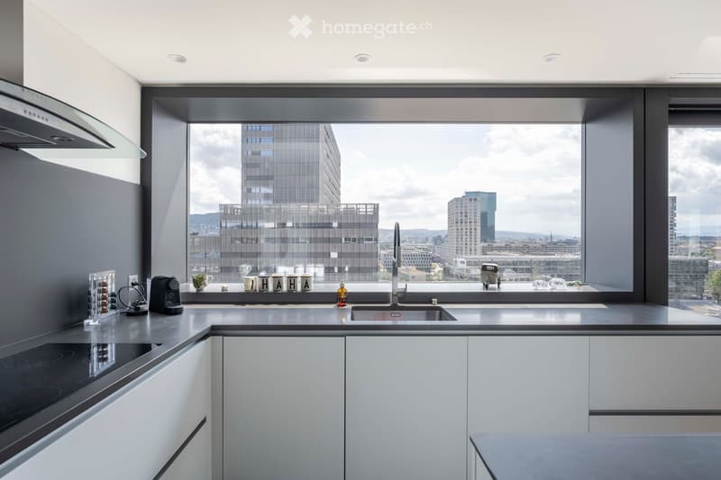 Modern kitchen with white cabinets, grey countertops, and large windows offering a cityscape view. Includes a sink, faucet, coffee maker, and induction stove.