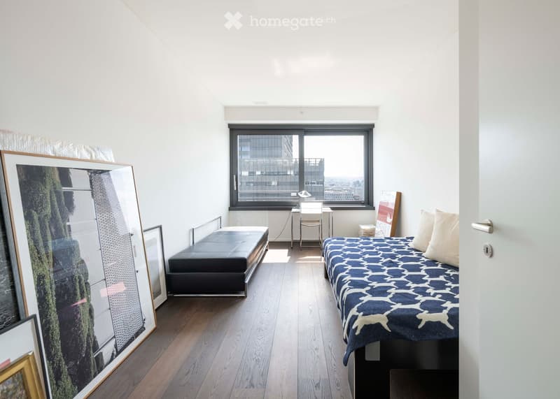 Modern bedroom with a large window offering a city view, featuring a bed with a blue and white patterned duvet, a bench, a desk with a chair, and several picture frames on the floor.