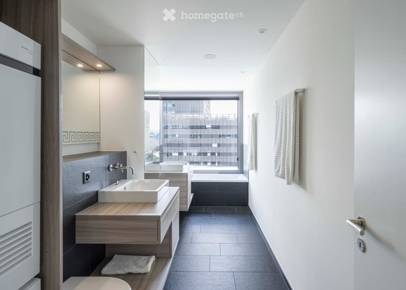 Modern bathroom with double sinks, a bathtub, a large window with city view, and contemporary fixtures.