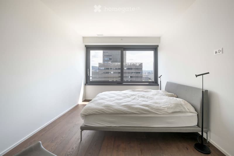Minimalist bedroom with a grey upholstered bed, white bedding, wooden flooring, and large window offering a city view.