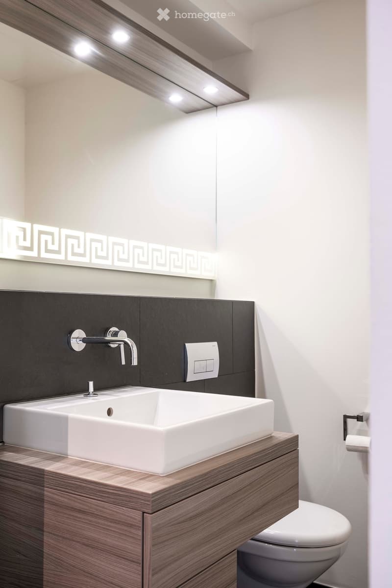 Modern bathroom with a white sink, dark wooden cabinet, chrome faucet, and a sleek mirror with decorative lighting. A toilet is visible in the background.