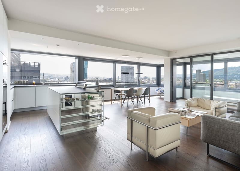 Modern open-plan living and dining area with large windows offering a city view. Features include a white leather sofa, a coffee table, a dining table with chairs, a kitchen island, and a glass display cabinet.