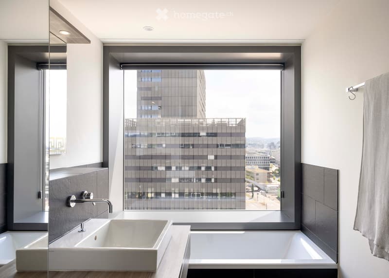 Modern bathroom with a large window offering a city view, featuring a white sink, bathtub, and a towel on the side.