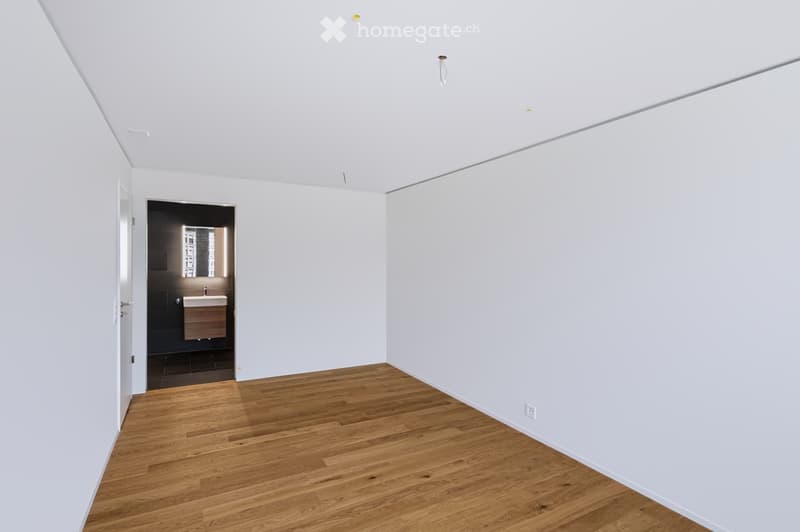 Empty room with white walls, wooden floor, and a small adjacent bathroom with a sink and mirror.