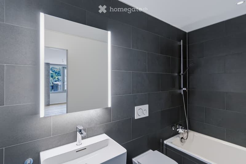 Modern bathroom with dark grey tiles, a large illuminated mirror, a white sink with a chrome faucet, a bathtub with a handheld showerhead, and a toilet.