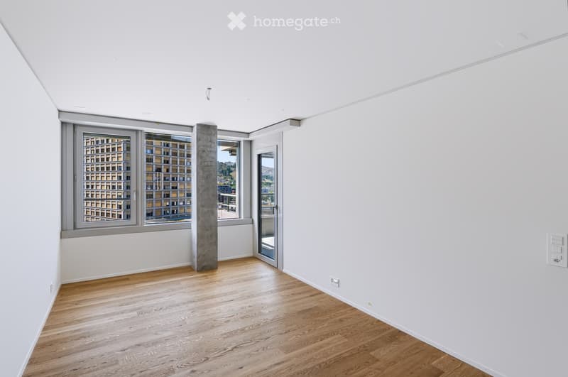 Empty room with wooden flooring, white walls, large windows, and a concrete pillar. No furniture or equipment present.