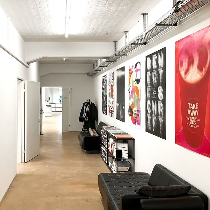 Modern hallway with white walls, wooden floor, black leather sofa, bookcase, coat rack, and several posters on the wall.