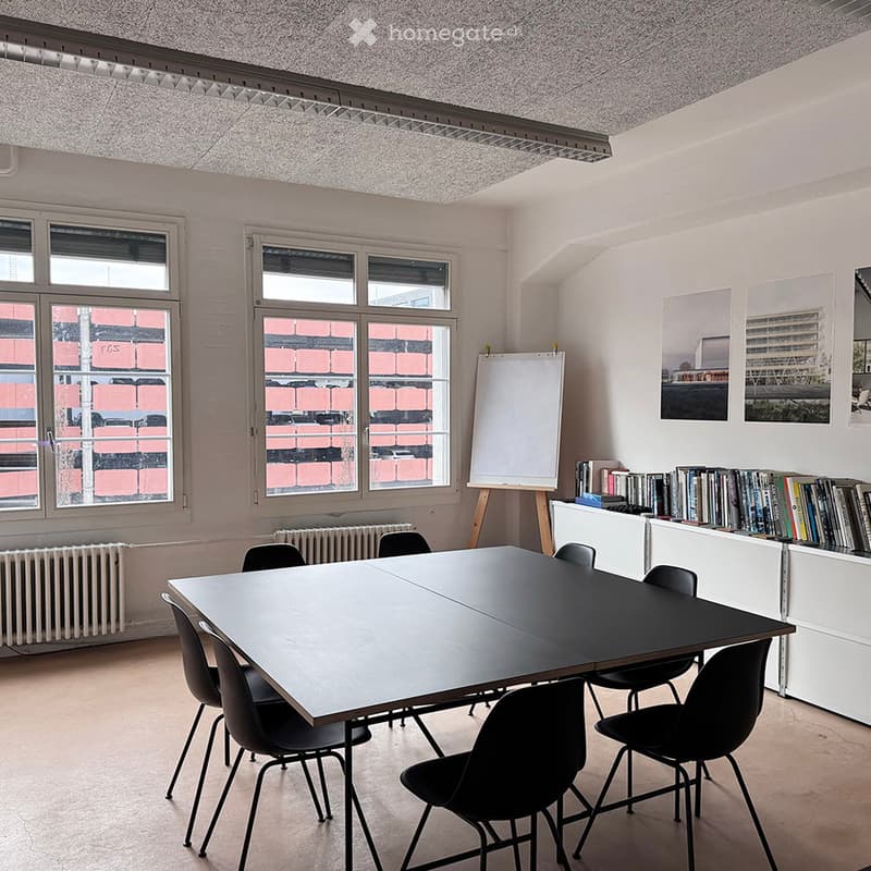 Bright room with large windows, a long black table surrounded by black chairs, a whiteboard on a stand, a bookshelf filled with books, and a radiator under the windows.