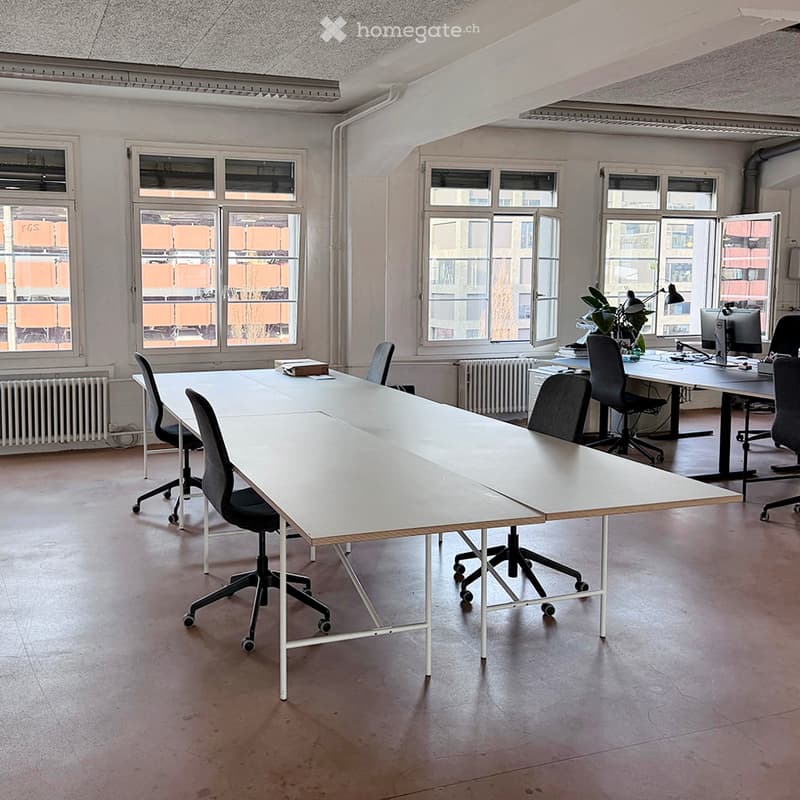 Bright office space with large windows, a long white conference table, several black office chairs, a computer setup with a monitor, and a small plant on a side table.
