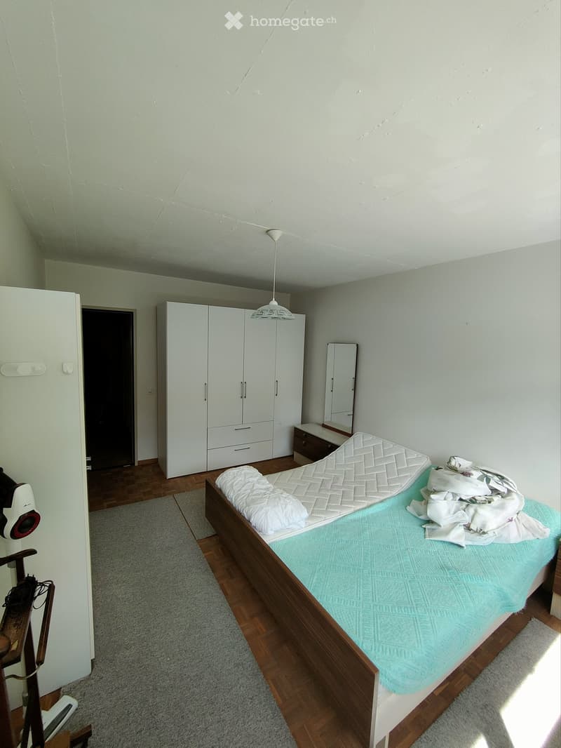A small bedroom with a wooden bed, white wardrobe, mirror, fan, and nightstand. The bed has a blue and white quilt and pillows.