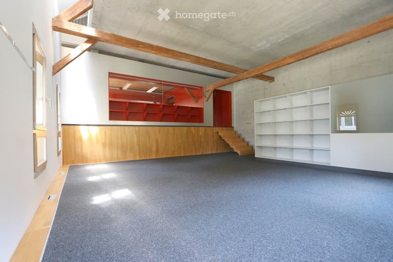 Modern, open-plan room with exposed wooden beams, grey carpet, white and red shelving units, and a wooden staircase leading to an upper level.