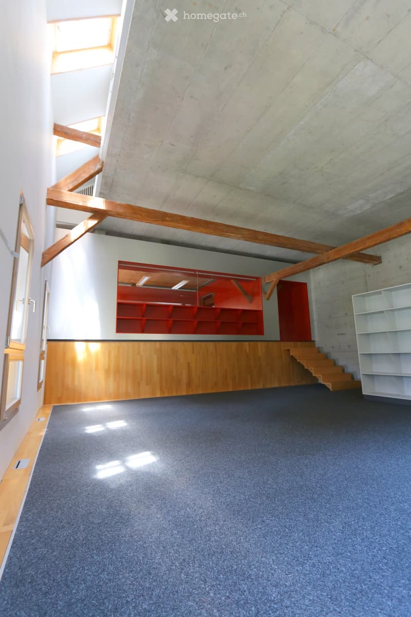 Empty room with blue carpet, wooden paneling, and exposed wooden beams. Features a staircase leading to an upper level and a red shelving unit in the background.