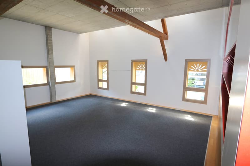 Empty room with white walls, wooden beams, and multiple windows, featuring a dark carpeted floor.