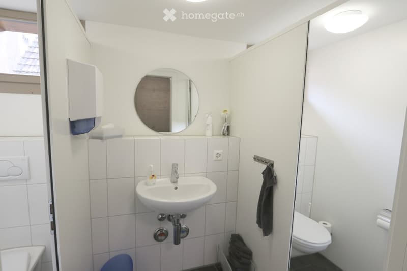 Modern, compact bathroom with white tiles, a round mirror, a sink, a toilet, and a towel hanging on a rack. A small window provides natural light.