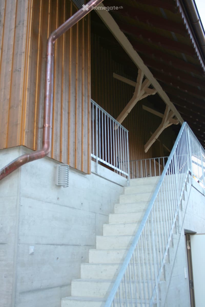 Concrete staircase with metal handrails leading up to a higher level, featuring a wooden ceiling and exposed pipes.