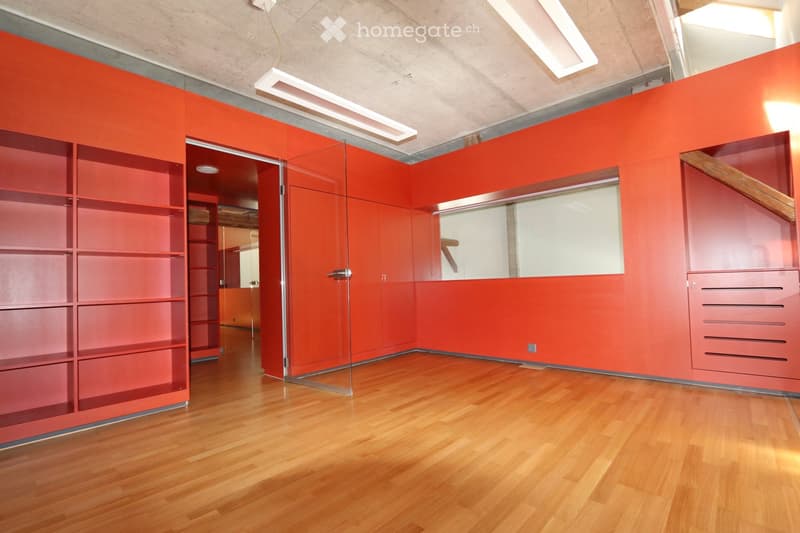 Modern unfurnished room with red walls, wooden flooring, built-in shelving units, and a glass partition leading to another space.