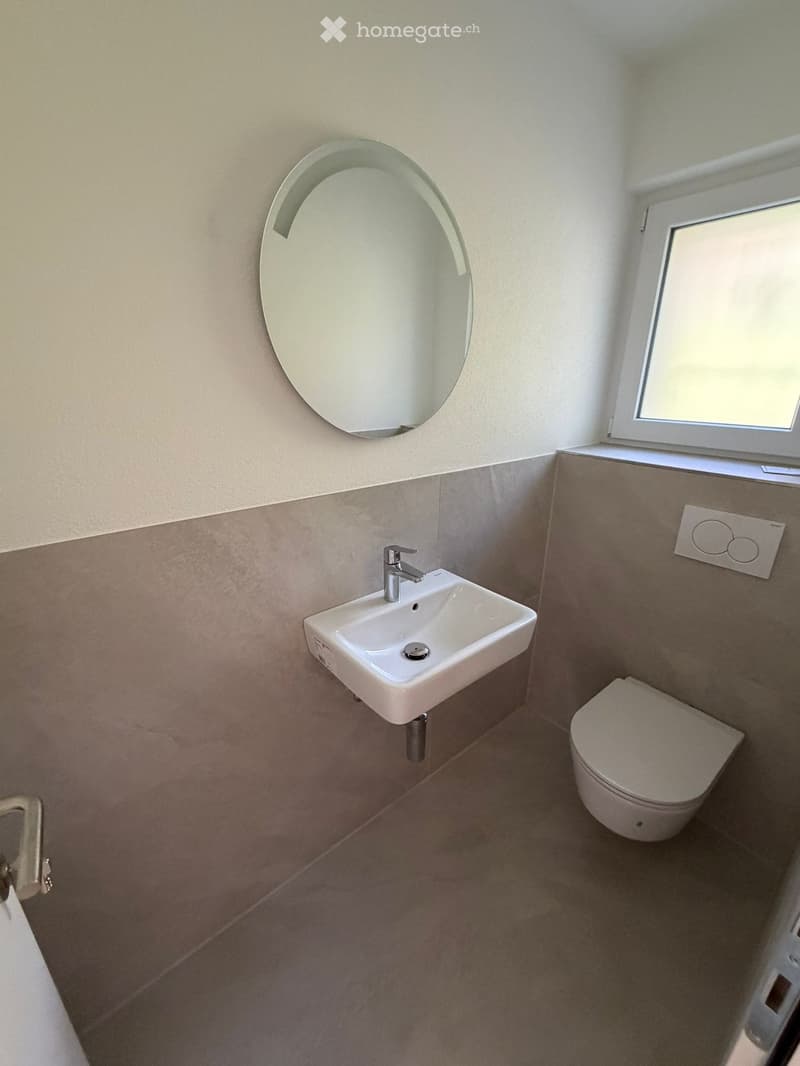 Modern bathroom with a white sink, chrome faucet, toilet, and a large round mirror above the sink. Light-colored tiles and a window provide natural light.