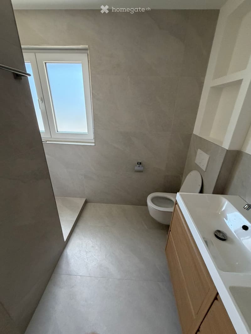 Modern bathroom with a toilet, sink, and wooden cabinet. The walls and floor are tiled in a neutral color scheme, and there is a window providing natural light.