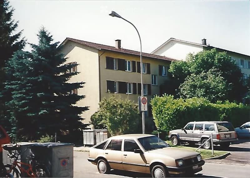 Two-story residential building with beige walls, multiple windows, and a tiled roof. The building is surrounded by trees and parked cars, with a streetlight and a stop sign visible in the foreground.
