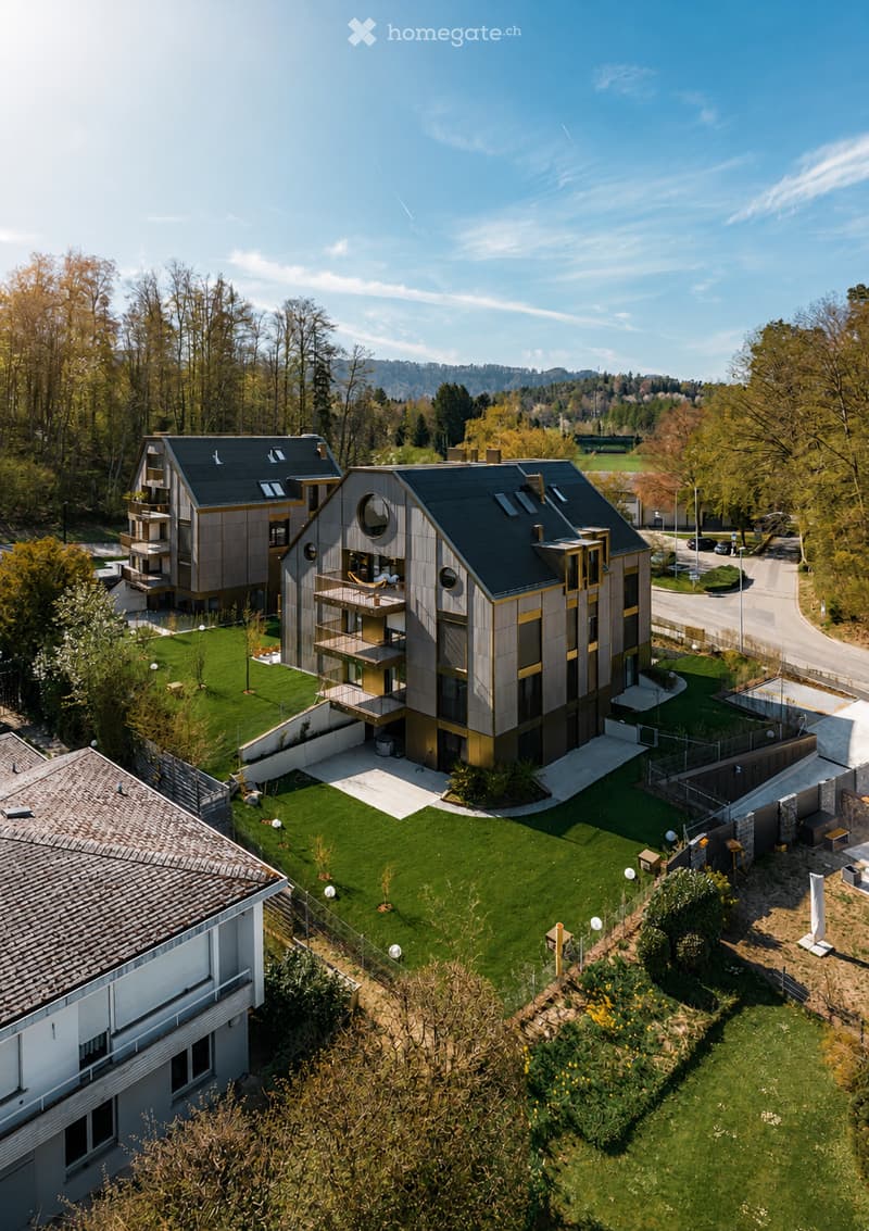 Aerial view of a modern residential complex with multiple buildings, surrounded by green lawns, trees, and a clear blue sky.