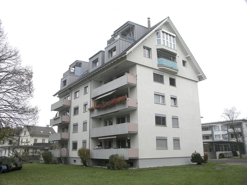 Modern multi-story apartment building with white facade, multiple balconies, and greenery in front.