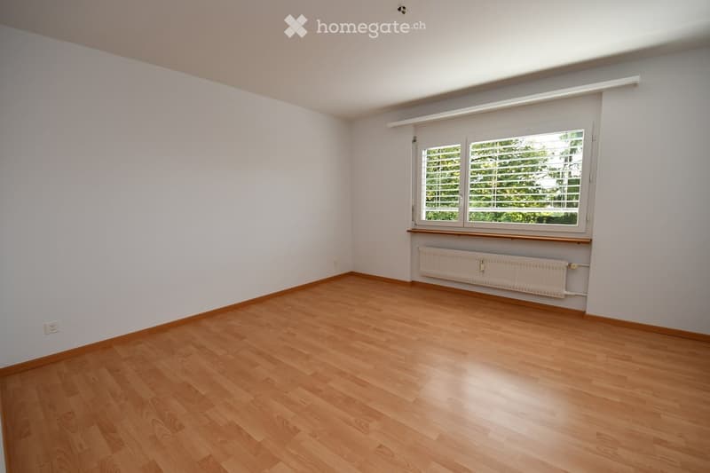 Empty room with wooden flooring, white walls, and a large window with blinds. A radiator is visible below the window.