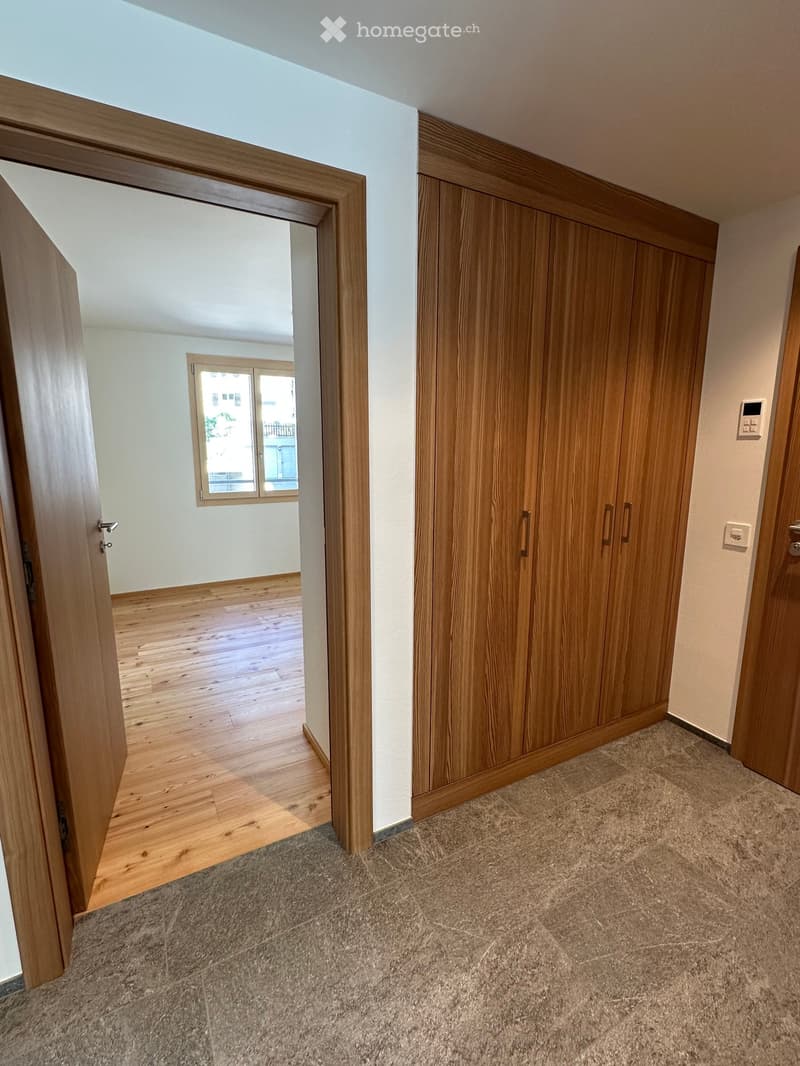 Empty hallway with wooden flooring, carpeted area, built-in wooden closet, and an open doorway leading to another room with wooden flooring and a window.