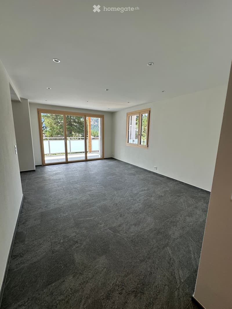 Empty room with grey tiled floor, white walls, recessed ceiling lights, large sliding glass door leading to balcony, and a window with wooden frame.