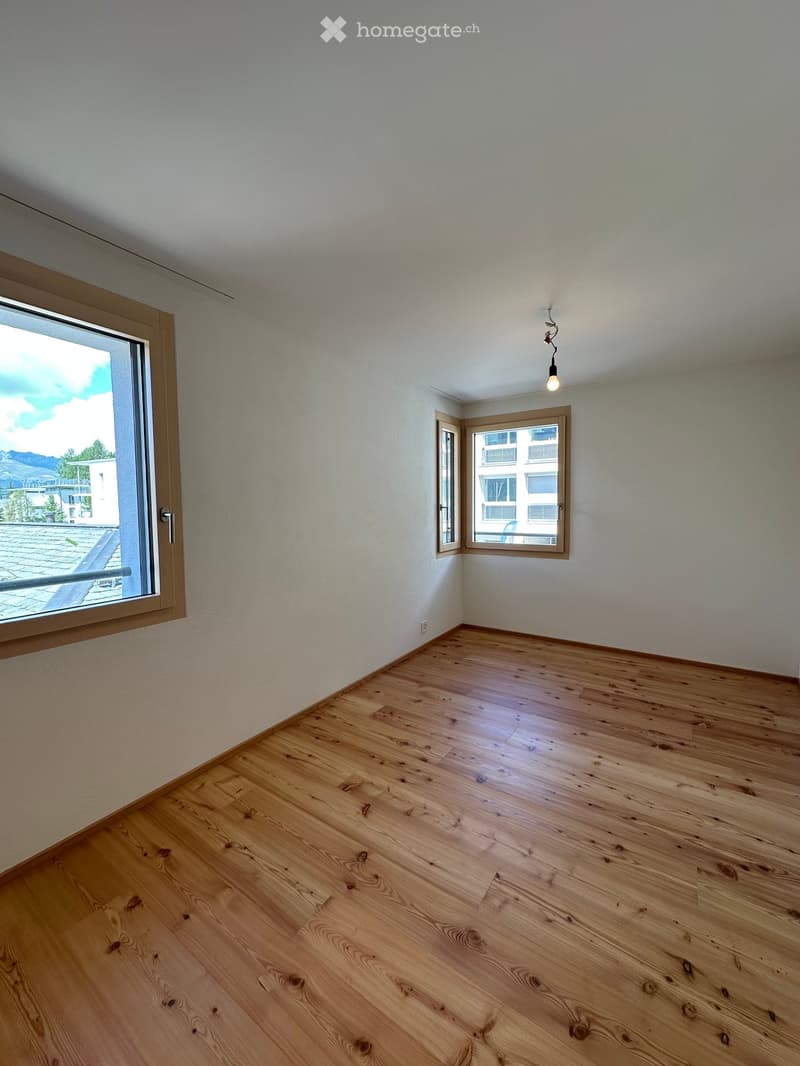 Empty room with wooden flooring, white walls, and large windows offering views of the outdoors.