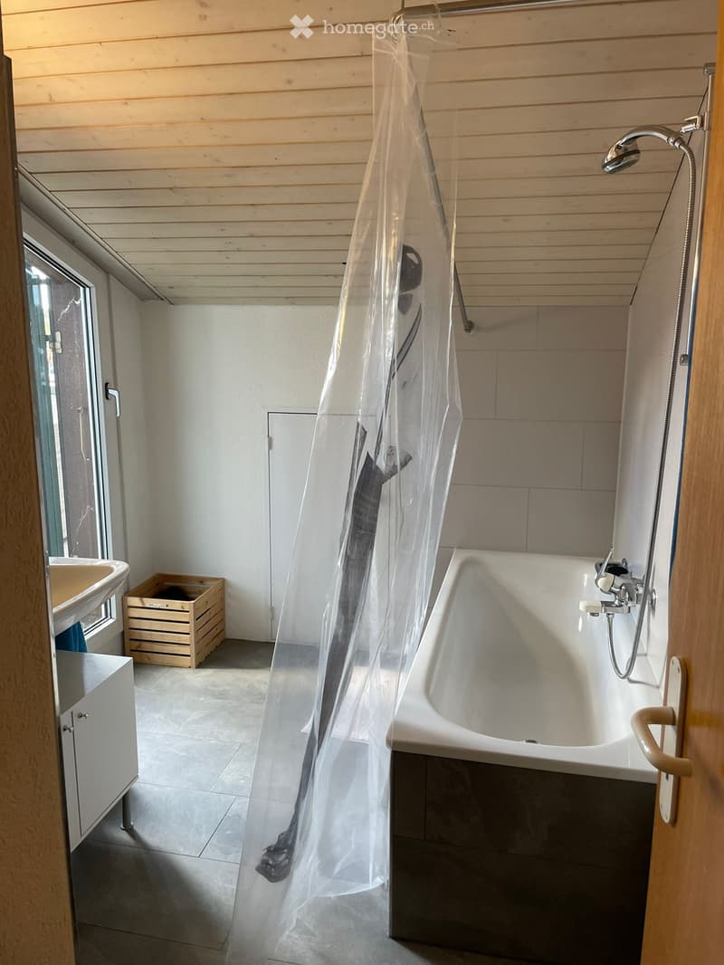 Modern bathroom with a freestanding bathtub, shower curtain, sink, and wooden cabinet. The space features light wooden paneling on the ceiling and walls, with a tiled floor.