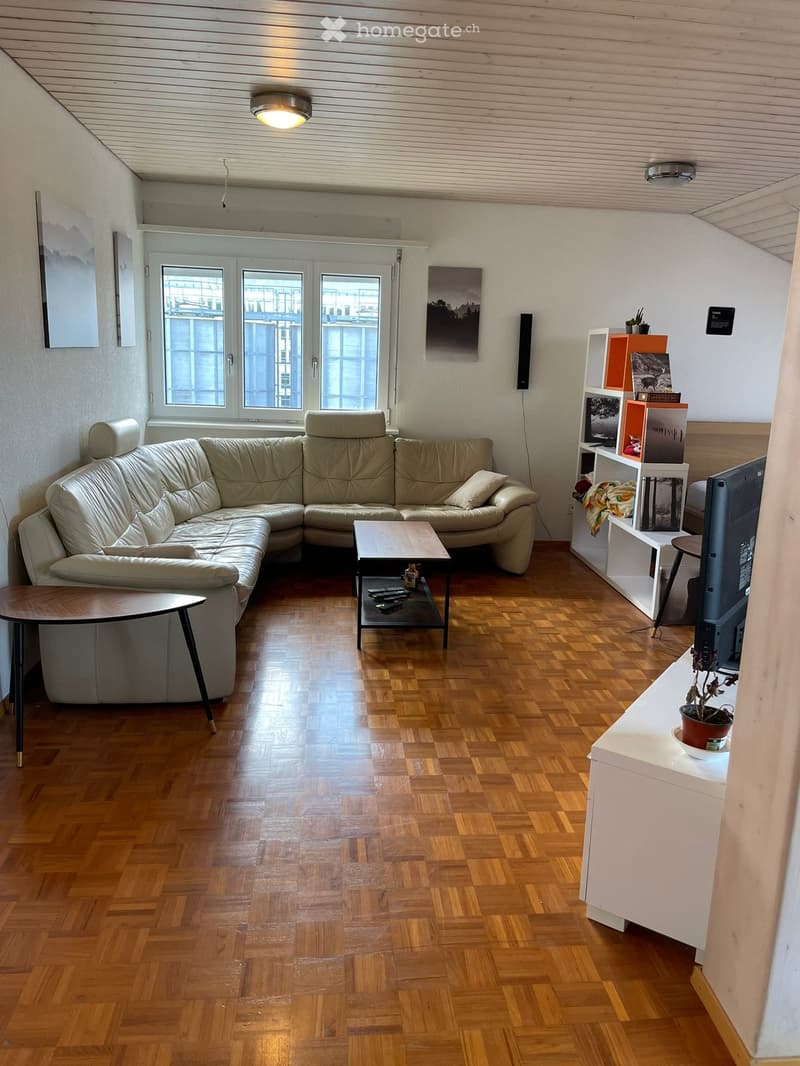 Modern living room with wooden flooring, large window, cream-colored sectional sofa, coffee table, small side table, shelf with books and decorative items, and a television on a stand.