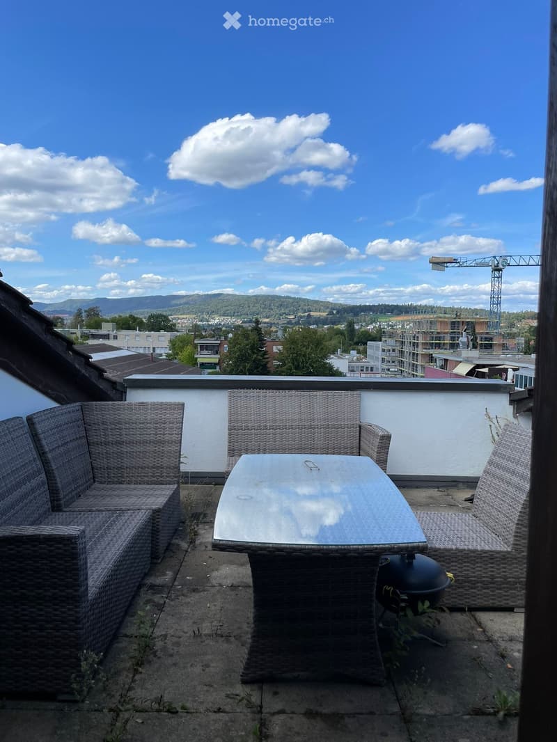 A rooftop terrace with a wicker sofa, glass table, and chairs, overlooking a cityscape with buildings and a crane in the distance.