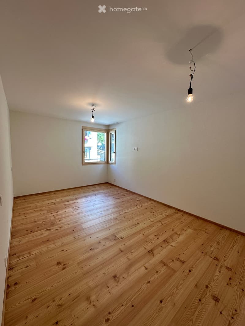 Empty room with wooden flooring, white walls, and a small window letting in natural light. The room has two ceiling lights and is completely unfurnished.