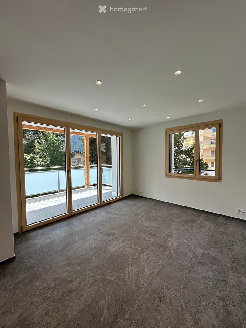 Empty room with grey tiled floor, white walls, recessed ceiling lights, large sliding glass door leading to a balcony, and a window with wooden frame.
