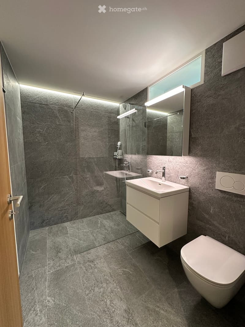 Modern bathroom with grey marble tiles, a white sink with a modern faucet, a toilet, a large mirror with built-in lighting, and a glass-enclosed shower area.