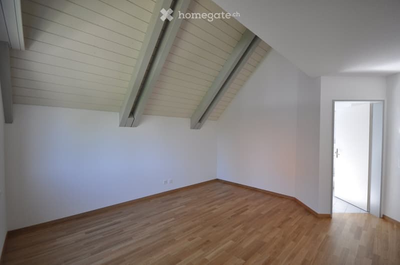 Empty room with wooden flooring, white walls, and exposed wooden beams on the ceiling. A doorway leads to another space.