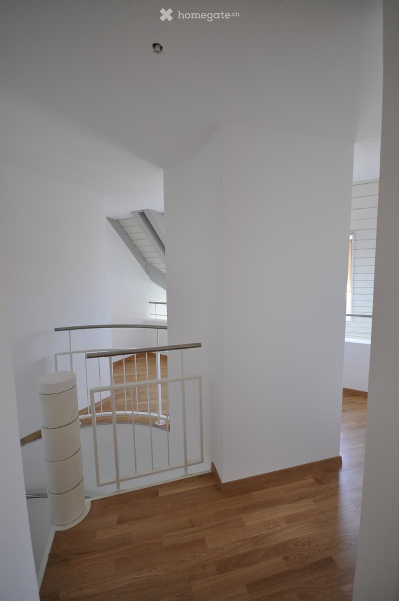 Spacious, modern hallway with wooden flooring, white walls, and a white railing leading to an upper level. Natural light streams in from the right side.