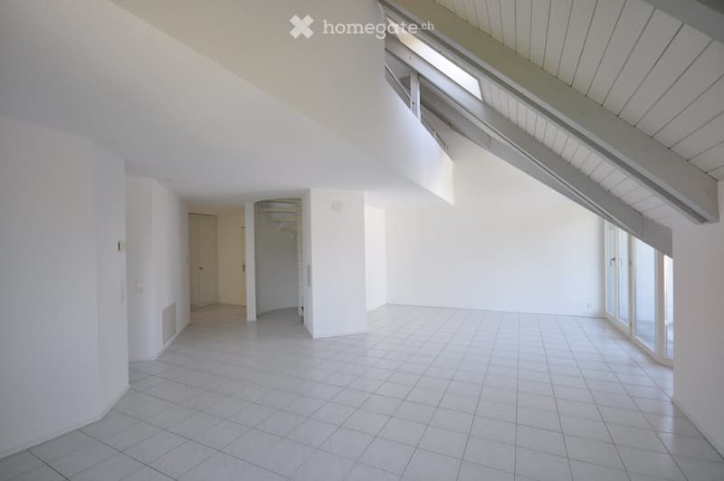 Empty room with white tiled flooring, white walls, and large windows allowing natural light. The room features a slanted ceiling with exposed wooden beams and a small closet area visible in the background.