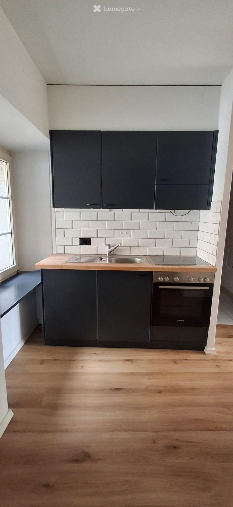 Modern kitchen with dark grey cabinets, white tiled backsplash, wooden countertop, sink, and built-in oven. Light wooden flooring throughout.