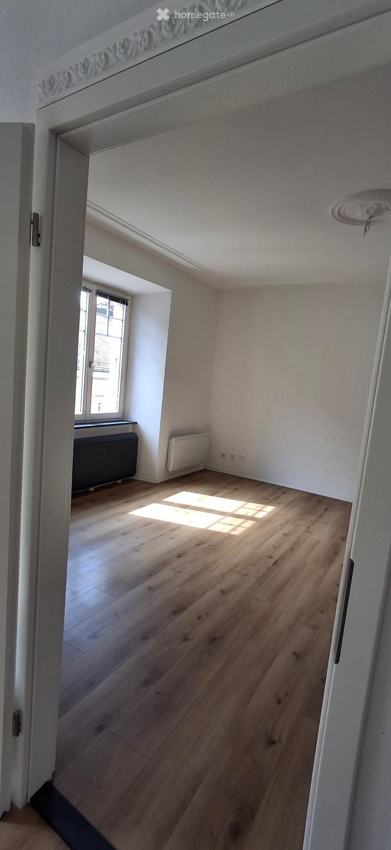 Empty room with wooden flooring, white walls, and a window letting in natural light. The room has a radiator and a door leading to another space.
