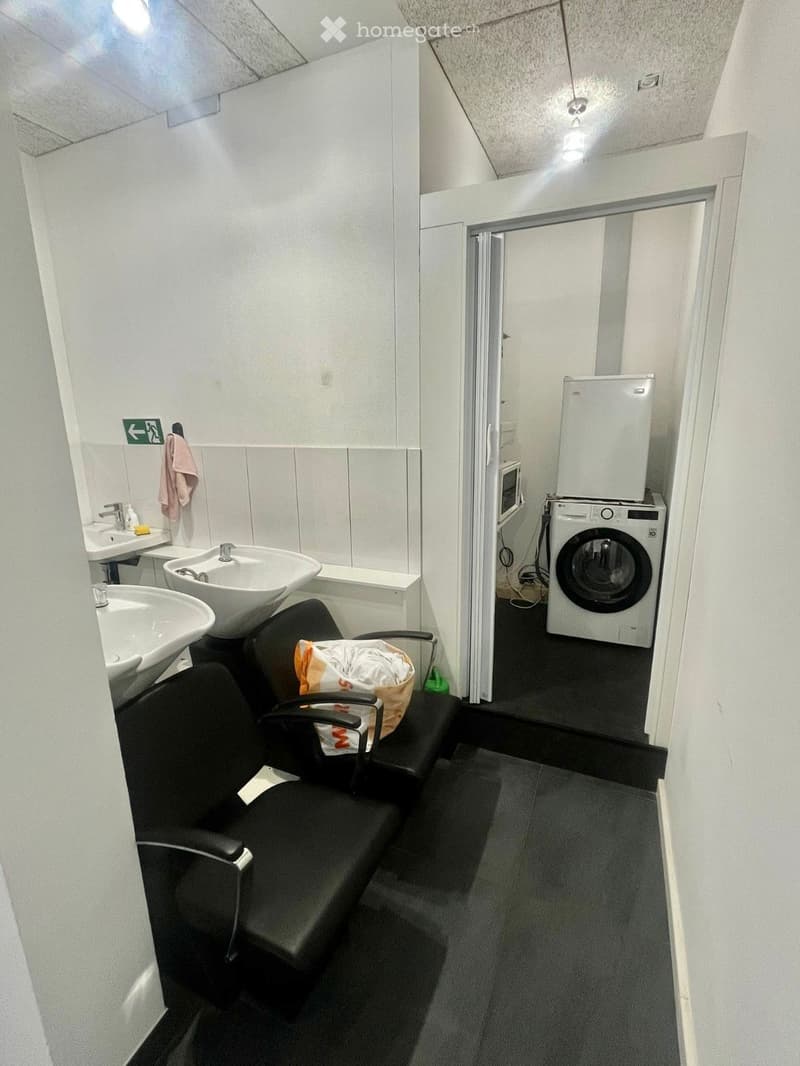 Bathroom with two sinks, a black chair, a washing machine, a refrigerator, and a microwave. A towel is hanging on the wall.