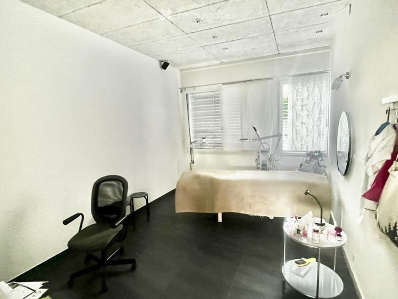 A small, well-lit room with a bed covered in a beige sheet, a black office chair, a small round table with medical supplies, a mirror on the wall, and a window with blinds.