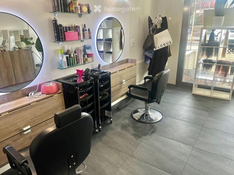 Modern salon with two black salon chairs, a wooden cabinet with shelves holding hair products, and a round mirror.