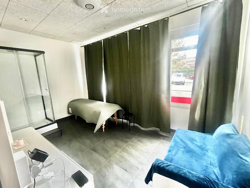 A small, well-lit room with a massage table, a stool, a chair, a table with a lamp, and a shower area with a glass enclosure.