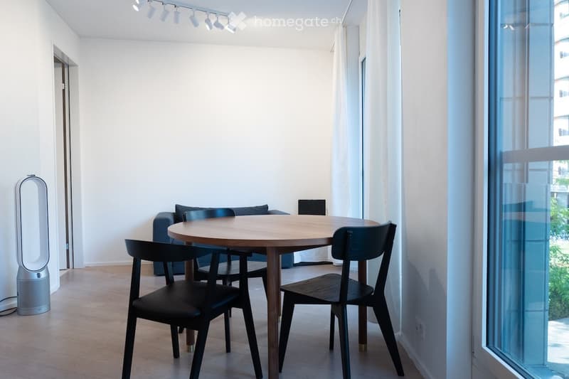 Modern, minimalist living space with a round wooden dining table, four black chairs, a grey sofa, and a sleek speaker on the floor. Large windows with sheer curtains allow natural light to fill the room.
