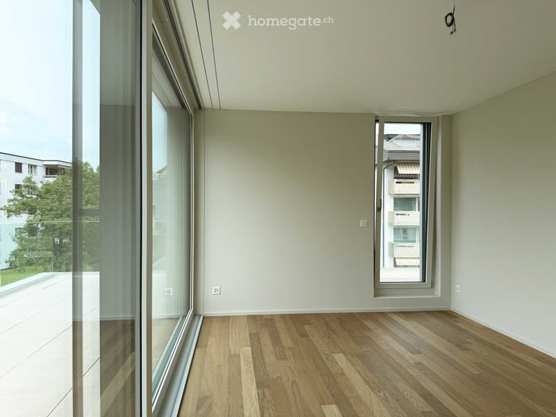 Empty room with light-colored walls, wooden flooring, and large windows offering views of the outside area.
