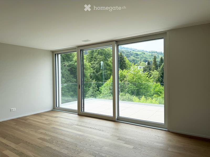 Empty room with wooden flooring, large sliding glass doors leading to a balcony with a scenic view of trees and hills.